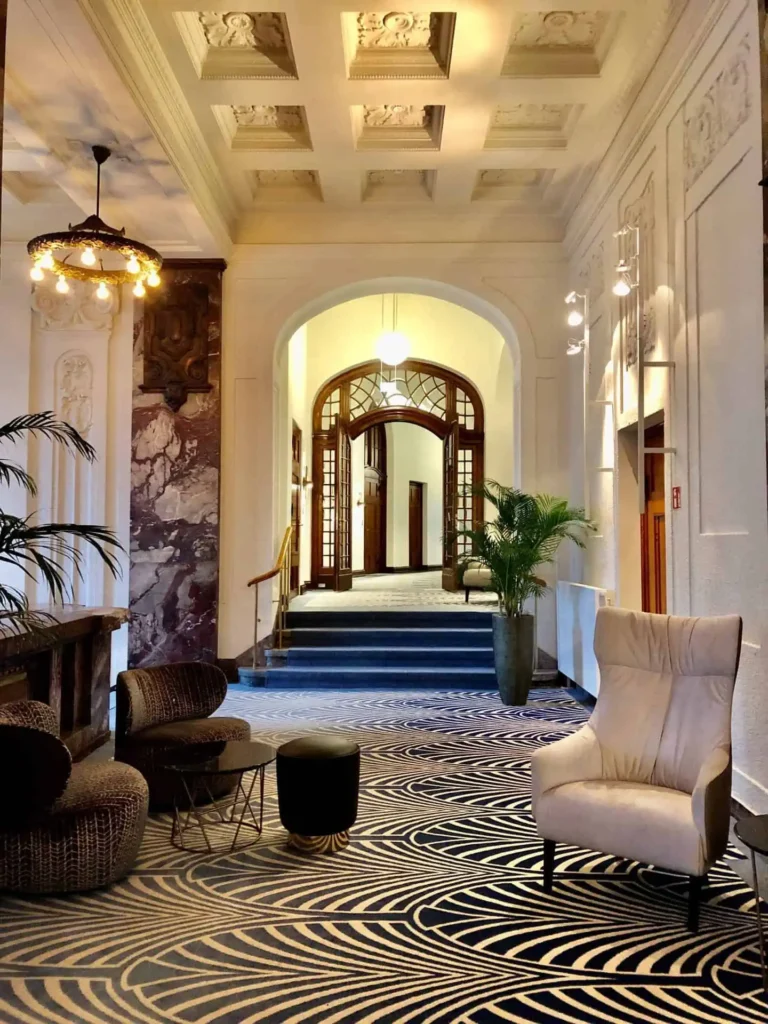 A stylish hotel lobby with patterned carpet, modern chairs, potted plants, and ornate ceiling details leads to a hallway with arched doorways and large windows. Warm lighting and nearby charging stations for electric cars add to the inviting atmosphere.