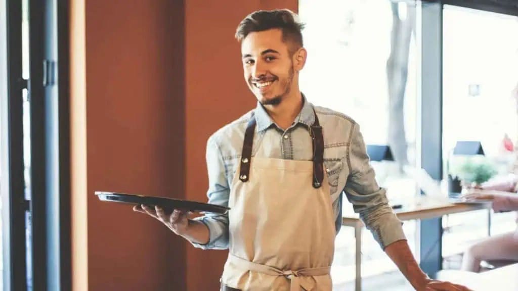 A smiling waiter wearing a light-colored apron and denim shirt holds a tray in one hand while standing inside a bright, modern café, reminiscent of the inviting spaces found in smart hotels with convenient charging stations.
