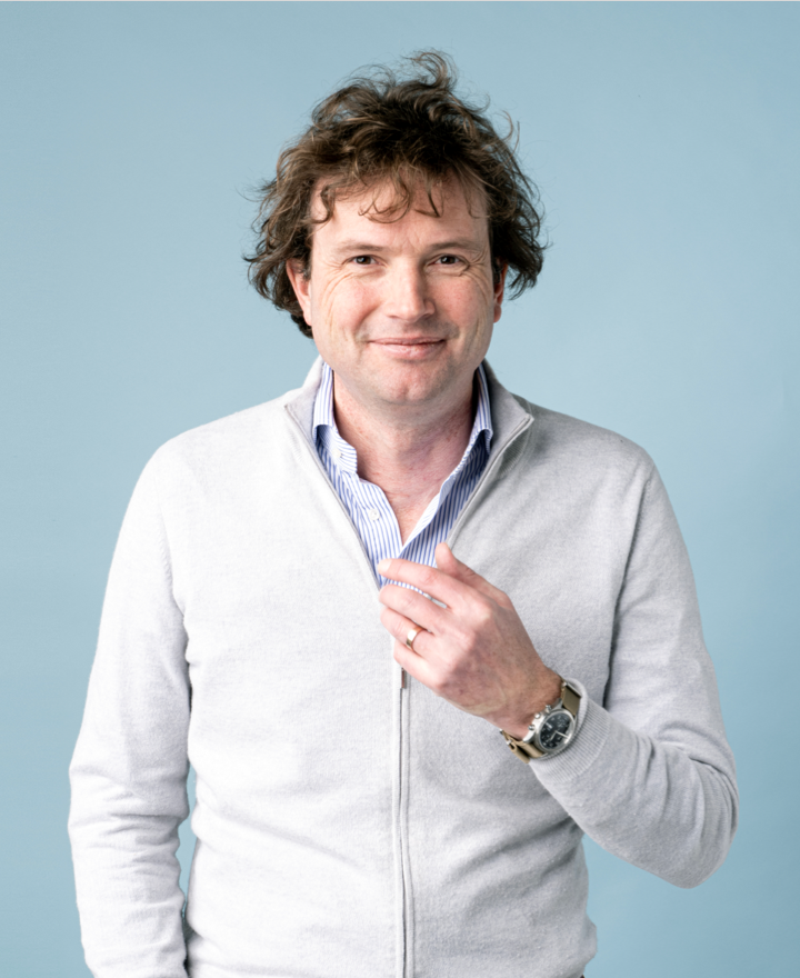 A man with wavy brown hair smiles at the camera, wearing a light grey zip-up sweater over a blue striped shirt. He stands by a plain light blue background, holding his sweater’s zipper—ready to talk about pluq’s free loading stations for charging.