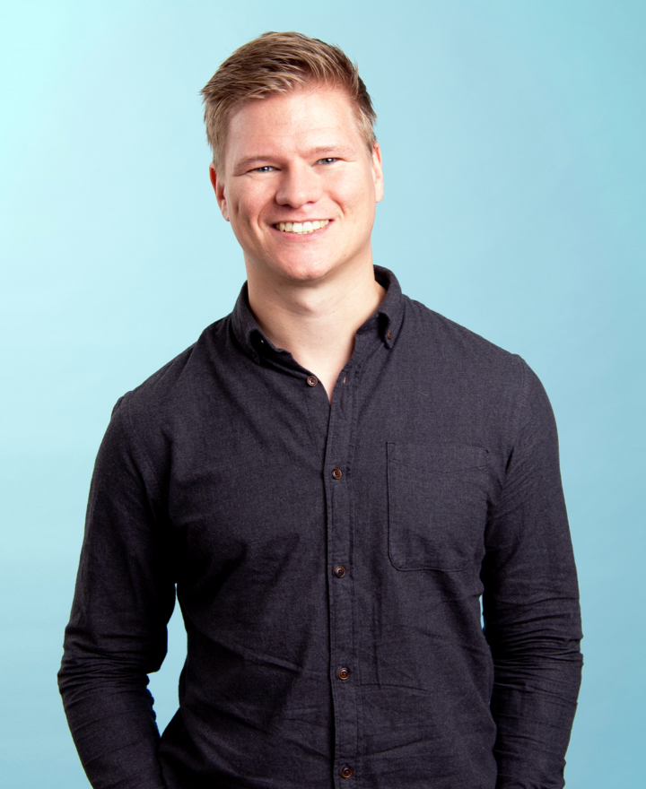 A smiling young man with short blond hair wearing a dark button-up shirt stands against a light blue background, holding an EV charge cable.
