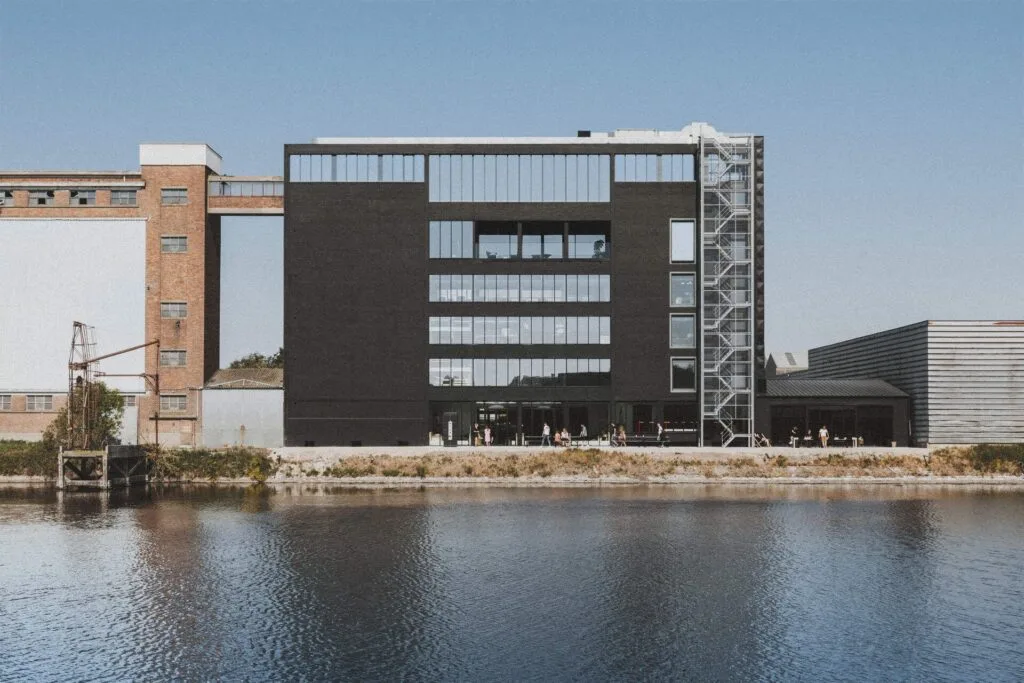 Een modern zwart gebouw met grote ramen staat aan het water, met rechts een metalen buitentrap. Bij de ingang verzamelen mensen zich bij oplaadstations van pluq, terwijl oudere industriële gebouwen op de achtergrond staan.