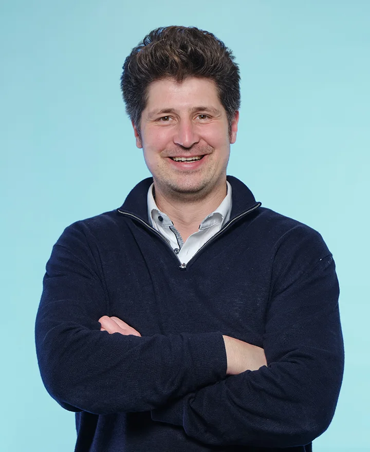 A man with short, dark hair smiles with his arms crossed, wearing a navy blue sweater over a collared shirt, standing against a solid light blue background—ready to talk about Pluq and its free loading stations for easy charging.