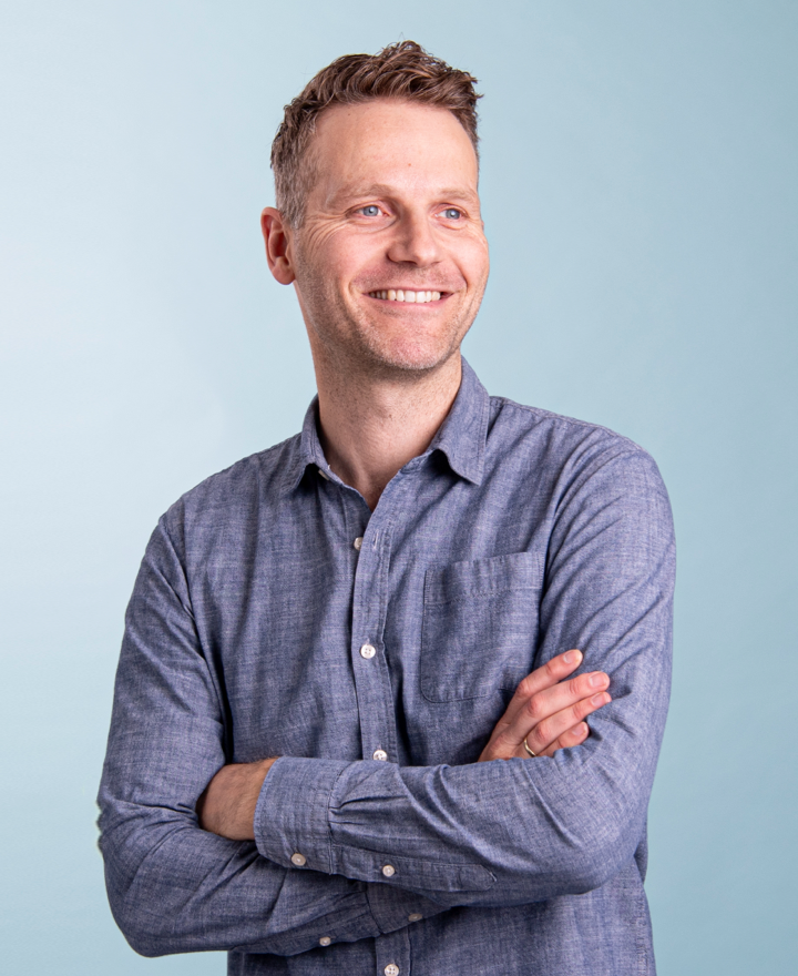 A man with short brown hair, wearing a blue button-up shirt, stands with arms crossed and smiles while looking to the side against a light blue background, evoking the modern spirit of electric cars.