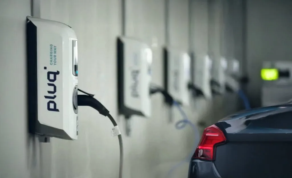 Eine Reihe von Ladestationen für Elektrofahrzeuge ist an einer Wand montiert, an der ein dunkel gefärbtes Auto lädt. Die pluq-Stationen bieten bequeme, kostenlose Ladestationen in einer überdachten Parkeinrichtung.