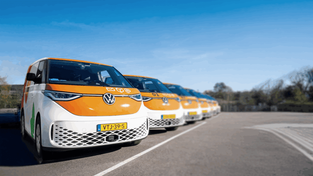 Une rangée de fourgonnettes Volkswagen orange et blanches, dont certaines sont électriques, est garée en file indienne avec des plaques d'immatriculation néerlandaises, par une journée ensoleillée, sous un ciel bleu limpide et avec des arbres en arrière-plan, probablement près de bornes de recharge gratuites.
