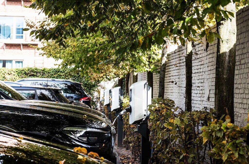 Electric cars are parked beside a white brick wall under leafy trees, connected to white electric vehicle charging stations. Some autumn leaves are scattered on the cars and bushes line the charging area.