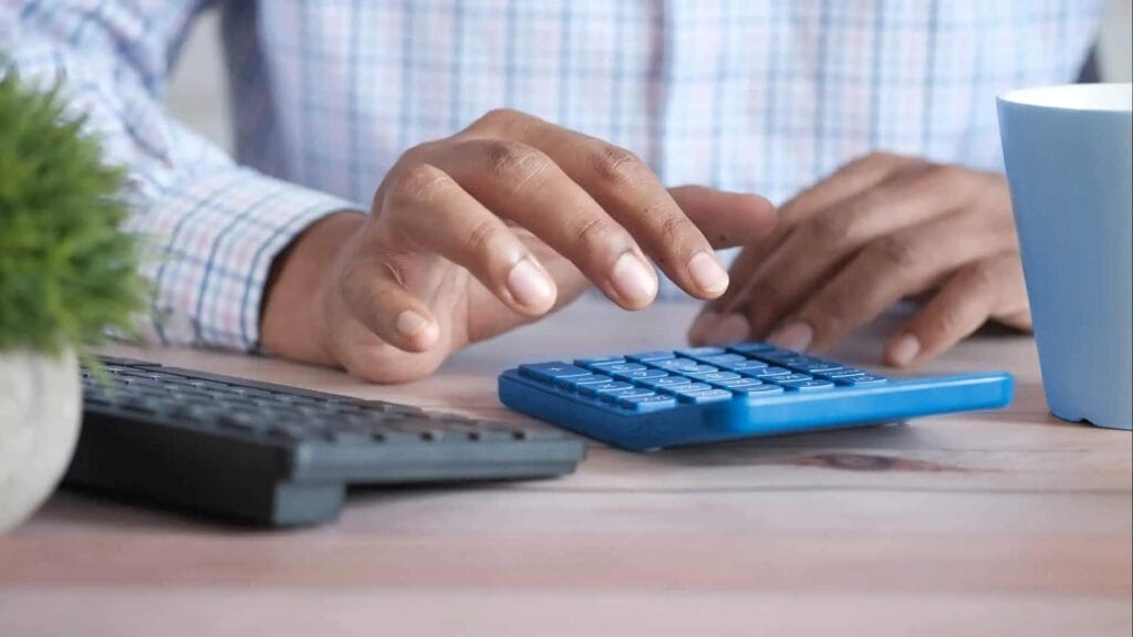 A person wearing a plaid shirt uses a blue calculator on a wooden desk. A black keyboard, a blue cup, and a small green plant are also visible on the desk.
