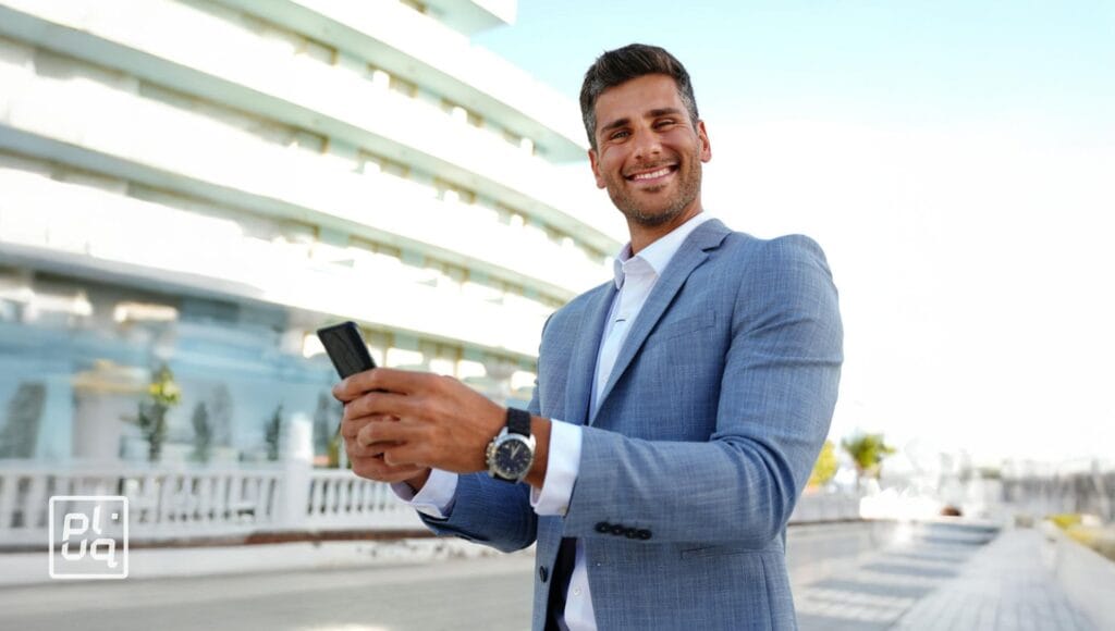 A man in a light blue suit smiles while using his smartphone outdoors in an urban setting, with a modern building in the background and sunlight illuminating the scene.