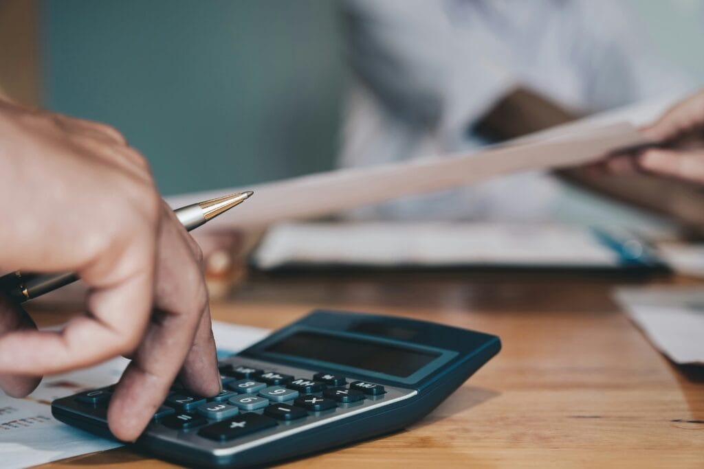 Close-up of a hand pressing calculator buttons with a pen, while another person holds papers nearby—both seated at a wooden desk, suggesting an accounting setting where financial questions or investment decisions are being considered.