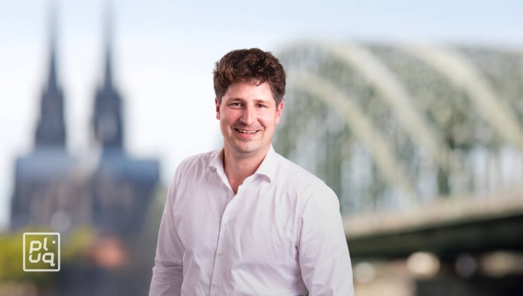 A smiling man in a white shirt stands in front of a blurred background featuring a large arched bridge and twin-spired cathedral. A white and yellow logo with the letters pluq appears in the lower left corner.