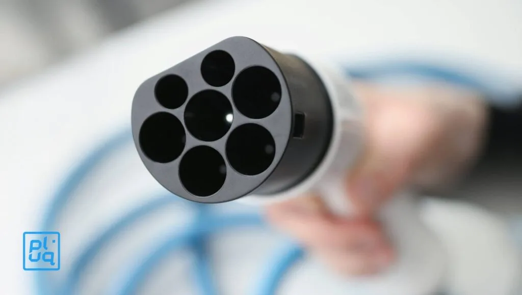 Eine Nahaufnahme einer Hand, die einen Ladestecker für Elektrofahrzeuge mit mehreren runden Anschlüssen und einem blauen Kabel im Hintergrund hält. Ein blaues pluq-Logo ist in der unteren linken Ecke zu sehen.