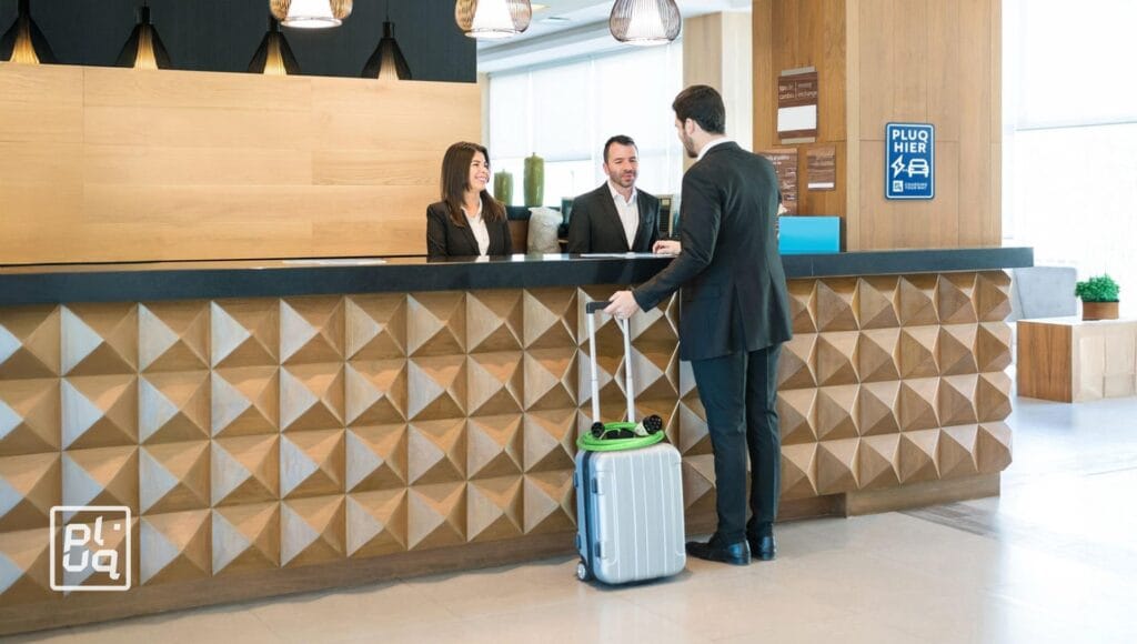 A man in a suit with a suitcase checks in at a modern hotel reception desk, speaking to two receptionists who are smiling behind the counter.