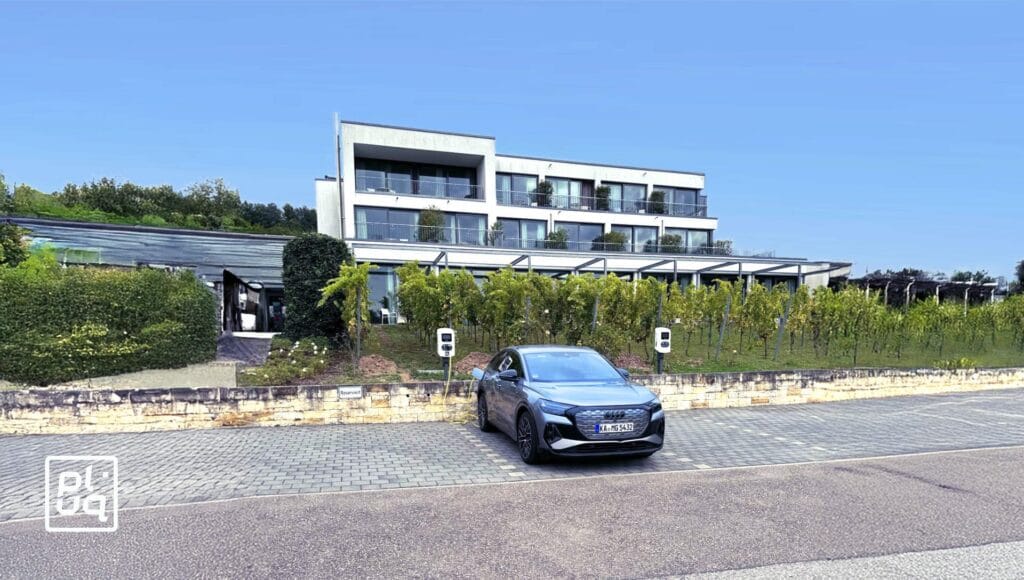 A black car is parked in front of a modern, multi-story building with glass balconies. The area includes greenery, electric vehicle charging stations, and a clear, sunny sky.