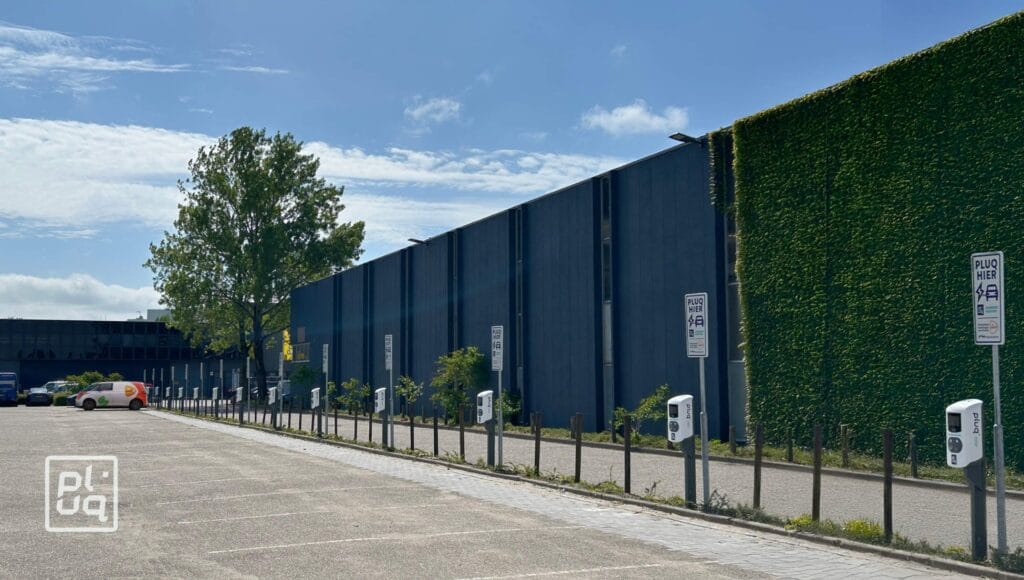 A mostly empty parking lot with several electric vehicle charging stations and signs. A large building with tall vertical windows and a green wall covered in plants is in the background under a sunny sky.