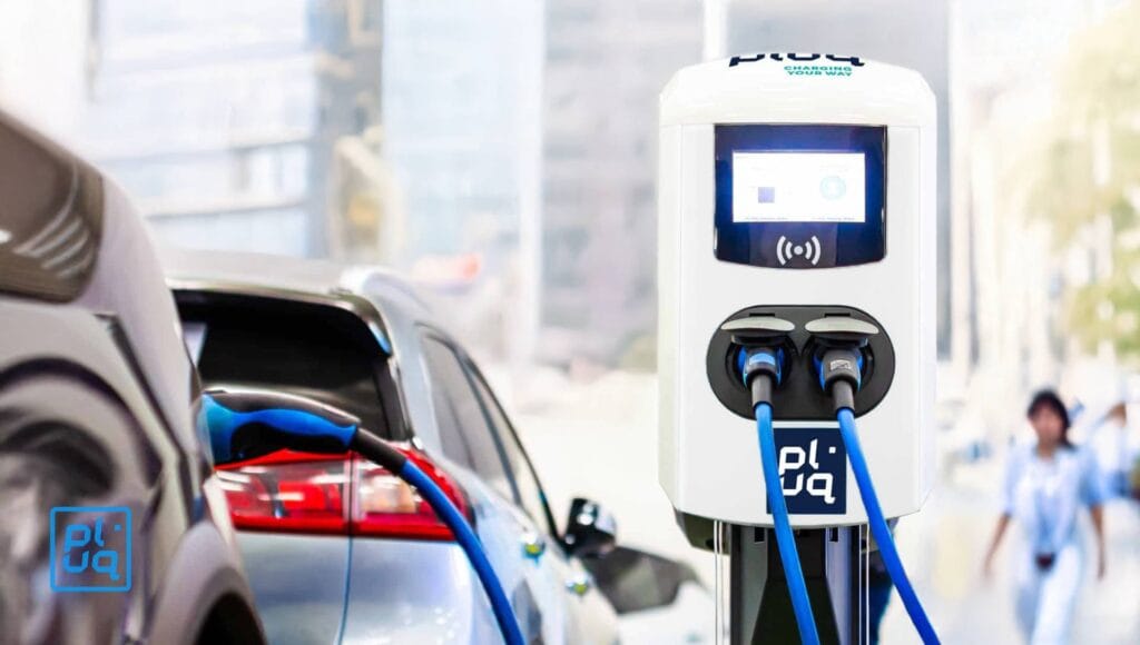 An electric car is plugged into a white charging station with a digital screen, set outdoors in an urban area with blurred buildings and people in the background.