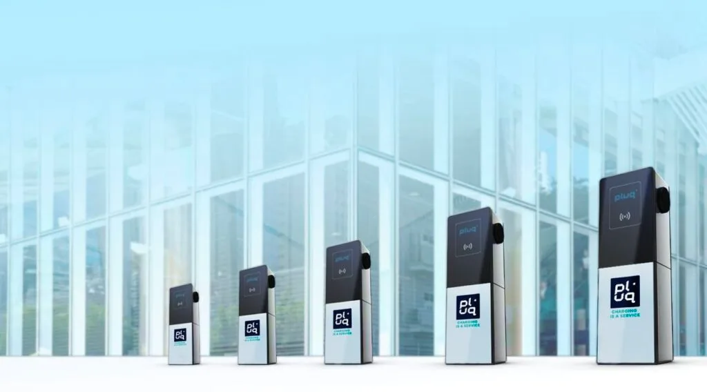 Eine Reihe von fünf Ladestationen für Elektrofahrzeuge ist im Freien vor einem modernen Glasgebäude aufgereiht, wobei jede Station mit dem pluq-Logo versehen ist.