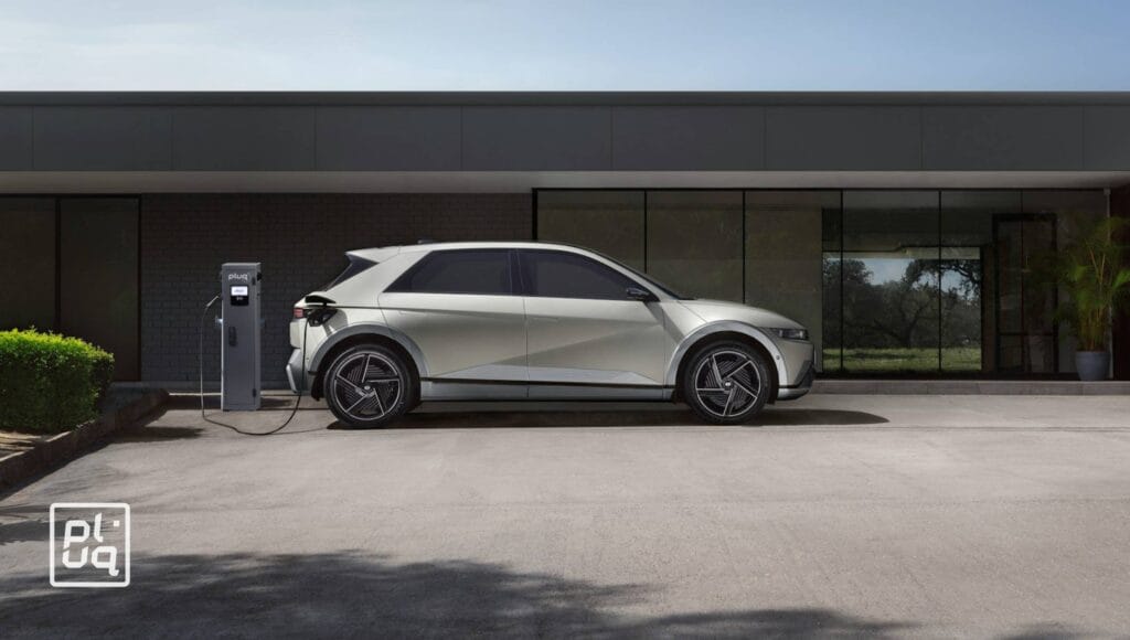 A silver electric car is parked and charging at a charging station in front of a modern building with large glass windows. Trees and plants are visible in the background.