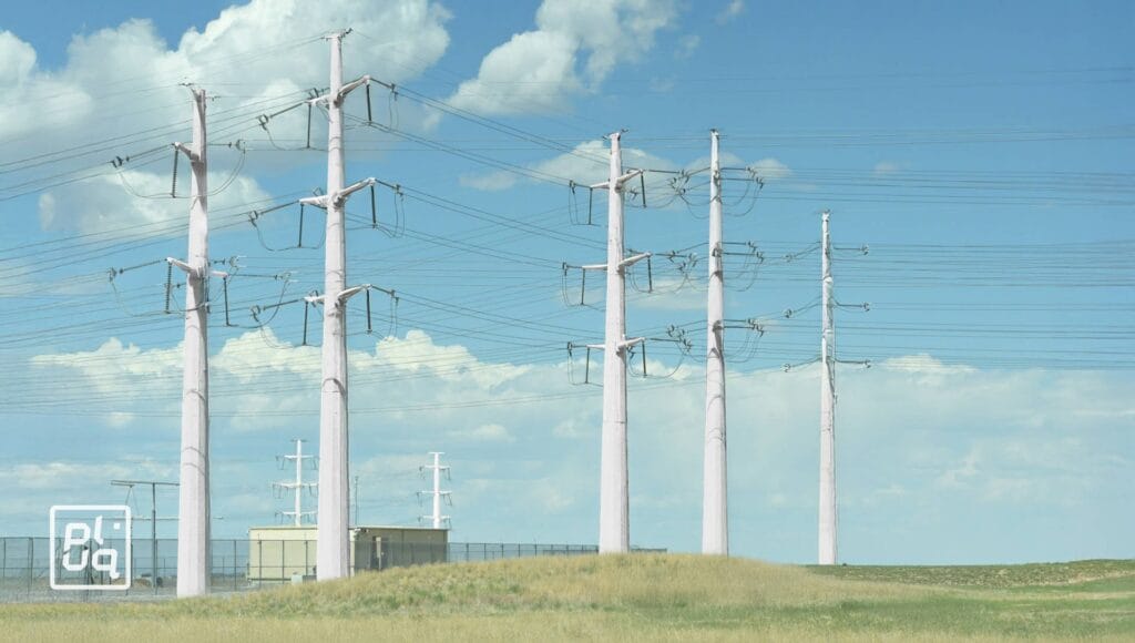 Rows of tall power lines stand in a grassy field under a blue sky with scattered clouds. A small utility building and a fenced area are visible near the base of the poles.