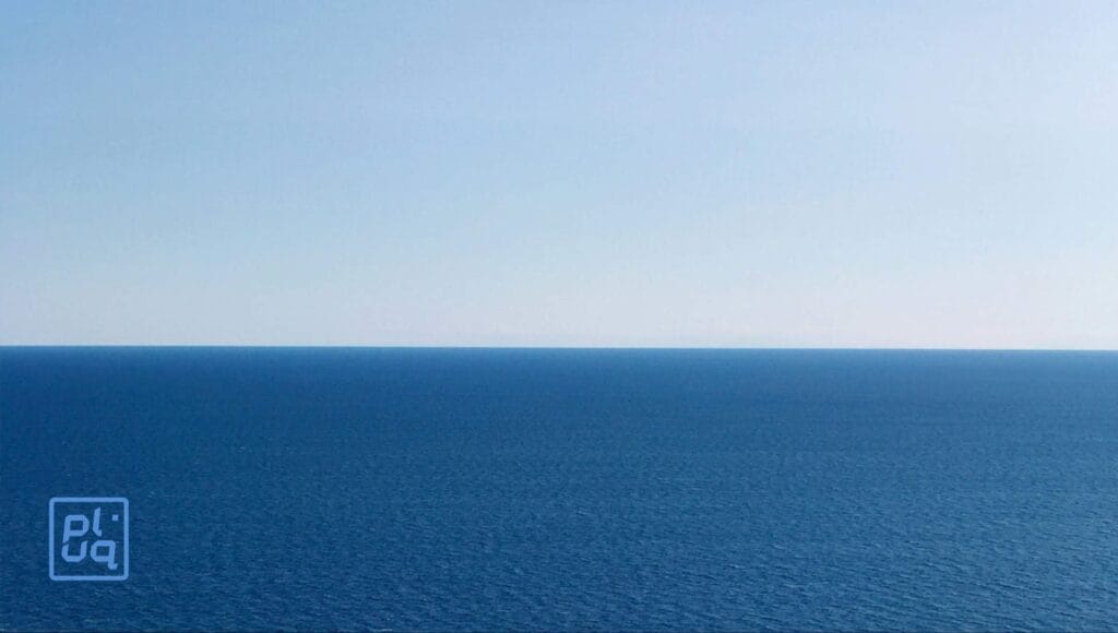 A clear blue sky meets a calm, deep blue ocean at the horizon. The water is gently rippled, and there are no visible boats, land, or clouds. A small, semi-transparent logo appears in the bottom left corner.
