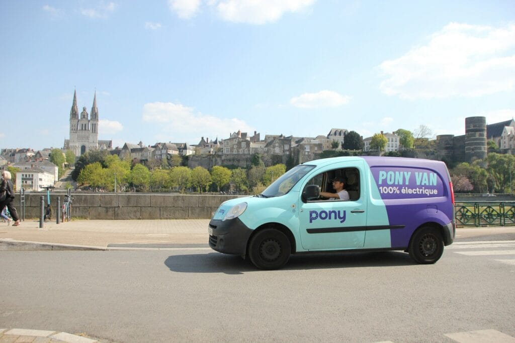 A light blue and purple electric van labeled PONY VAN 100% électrique drives along a riverside road in a European city, with historic buildings and a cathedral visible in the background under a blue sky.