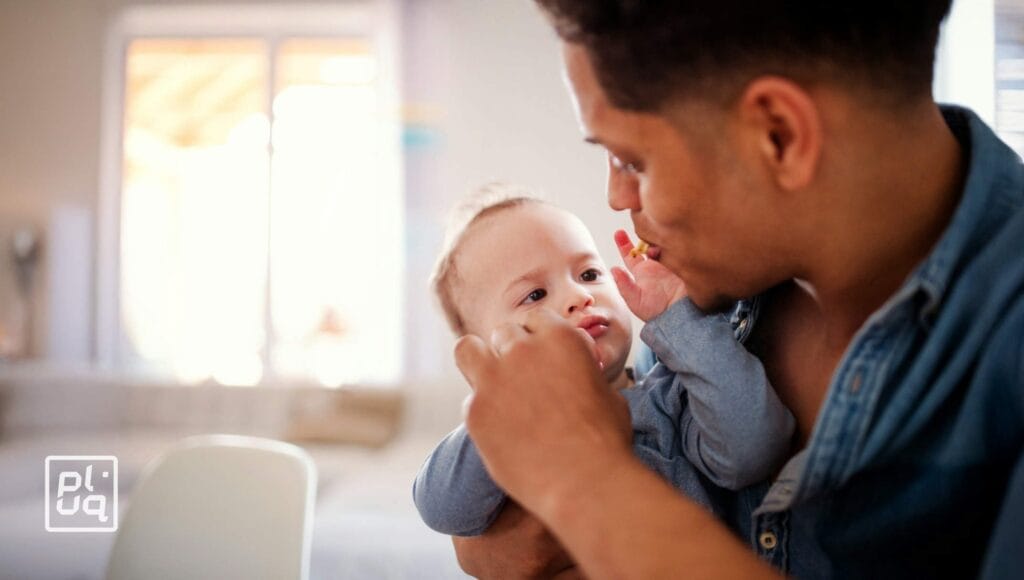 A man lovingly feeds a baby, holding the child in his arms while offering a spoonful of food. The baby looks up at the man in a bright, cozy room.