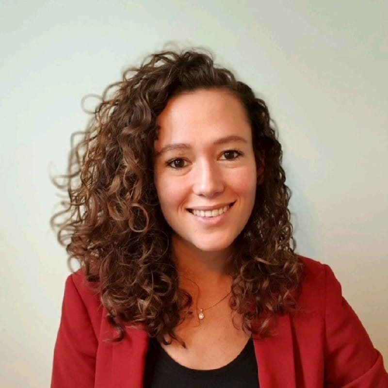 A woman with curly brown hair, wearing a red blazer over a black top, smiles at the camera in front of a light-colored background, making it look easy to capture a perfect moment in no time.
