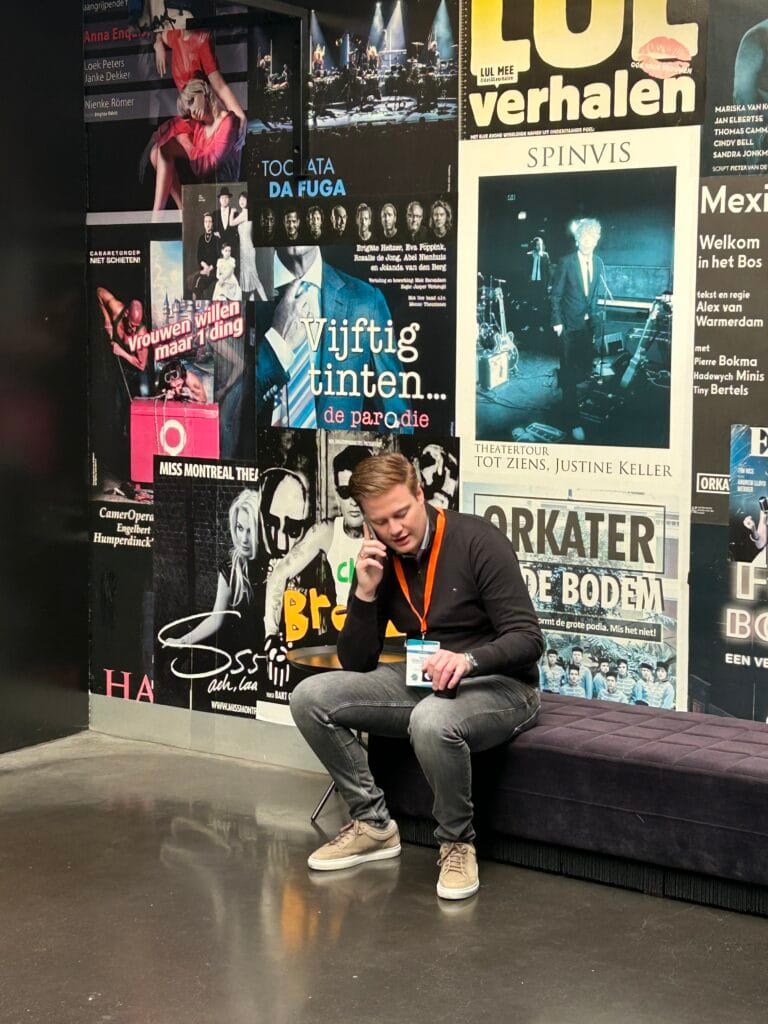 A man with a lanyard sits on a bench, talking on the phone in front of a wall covered with colorful posters for various events and performances.