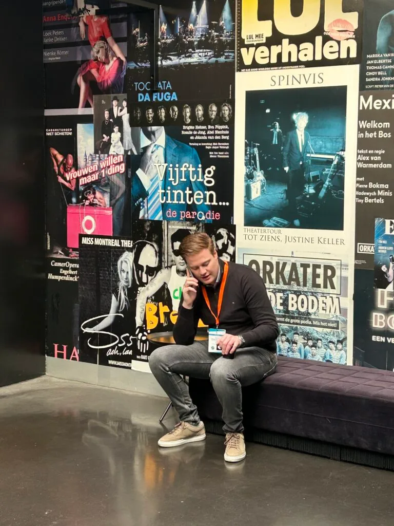 Ein Mann mit einem Schlüsselband sitzt auf einer Bank und telefoniert vor einer Wand, die mit bunten Plakaten für verschiedene Veranstaltungen und Aufführungen bedeckt ist.