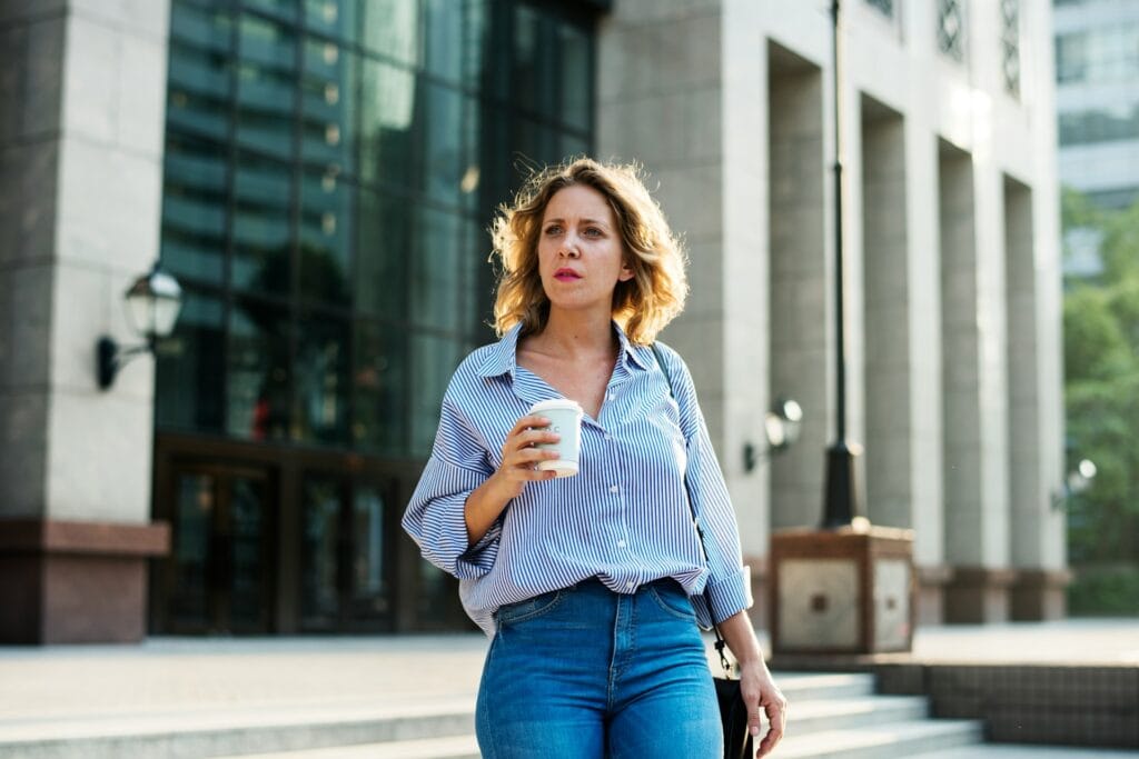 A woman with short blonde hair, wearing a blue striped shirt and jeans, holds a coffee cup while walking outside a modern building with large glass windows and columns.