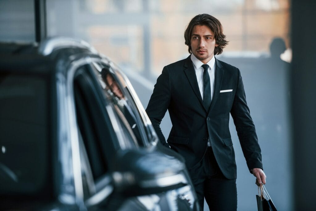 A man in a black suit and tie stands next to a car, holding a bag in his hand. He has shoulder-length brown hair and a serious expression, with a blurred indoor background.