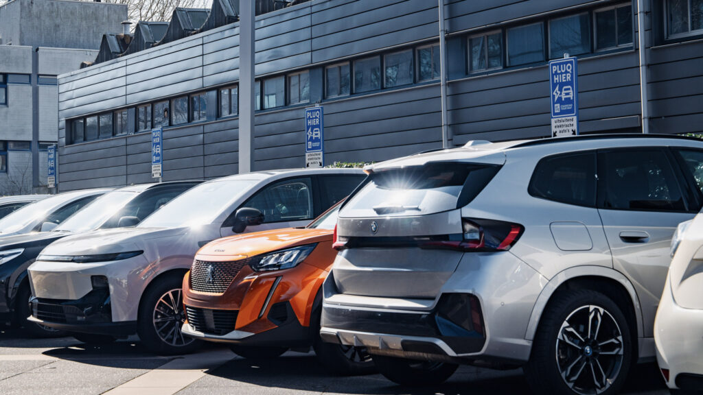 Eine Reihe geparkter Elektrofahrzeuge vor einem modernen Gebäude, hinter den Autos sind Schilder mit der Aufschrift „Plug Here“ für EV-Ladestationen zu sehen.