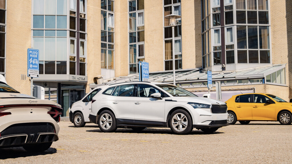 Plusieurs voitures, dont un SUV blanc, sont garées devant un immeuble moderne doté de grandes baies vitrées. Des panneaux bleus portant des inscriptions blanches sont placés près des places de stationnement. La lumière vive du soleil projette des ombres sur le sol.