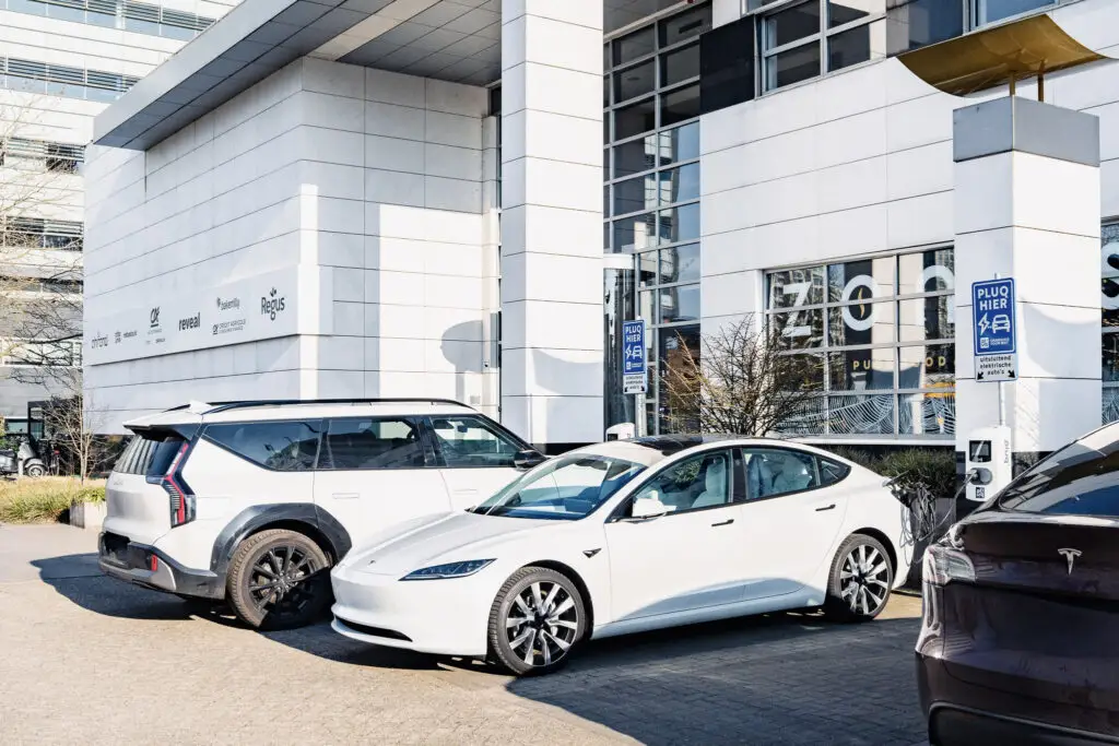 Two white electric cars are parked at charging stations outside a modern office building with large windows and company signs on the wall.