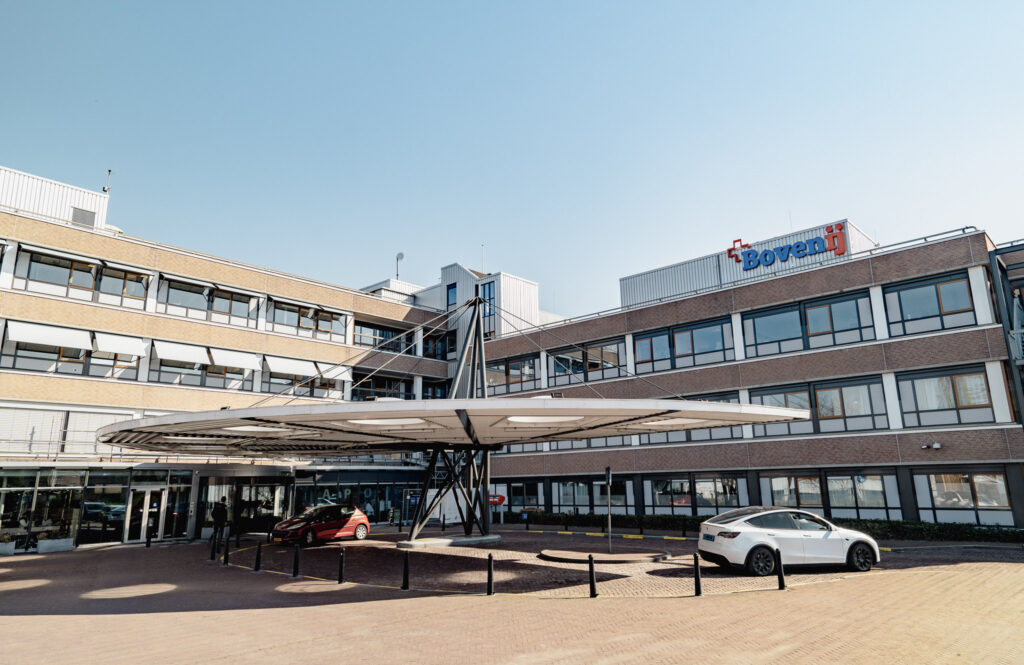 Modernes Krankenhausgebäude mit großen Fenstern und einem runden Eingangsvordach. Vor dem Gebäude stehen zwei Autos, und an der oberen Ecke des Gebäudes ist ein Schild mit der Aufschrift „Bovenij“ zu sehen. Über dem Gebäude erstreckt sich ein strahlend blauer Himmel.