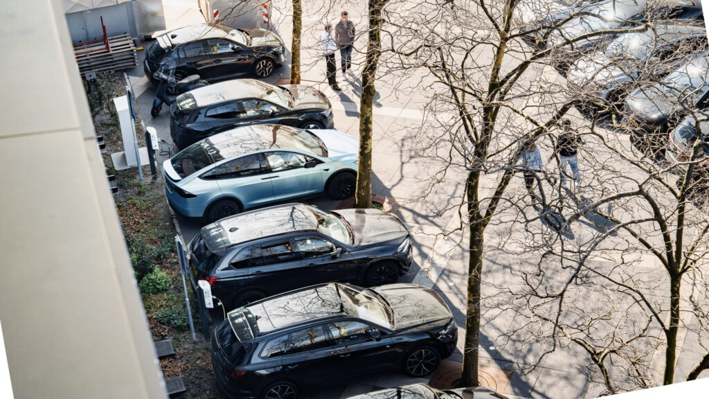 Vue aérienne de plusieurs voitures garées, dont une voiture bleu clair au milieu d'une rangée de véhicules principalement noirs, le long d'une rue bordée d'arbres dénudés. Une personne marche sur le trottoir à proximité.