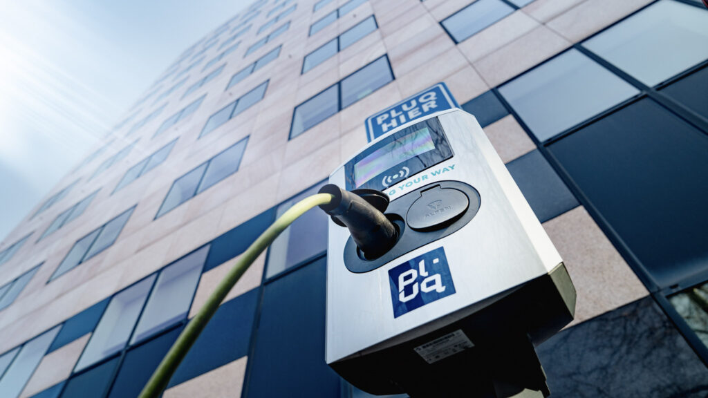 Gros plan sur une borne de recharge pour véhicule électrique avec un câble branché, avec en arrière-plan un immeuble de bureaux moderne en verre et en pierre. La perspective est orientée vers le haut, en direction du ciel.