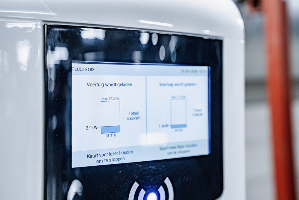 Close-up of a digital charging station screen displaying information in Dutch about a vehicle charging session, including power usage and duration, with part of the white station and a blurred background visible.