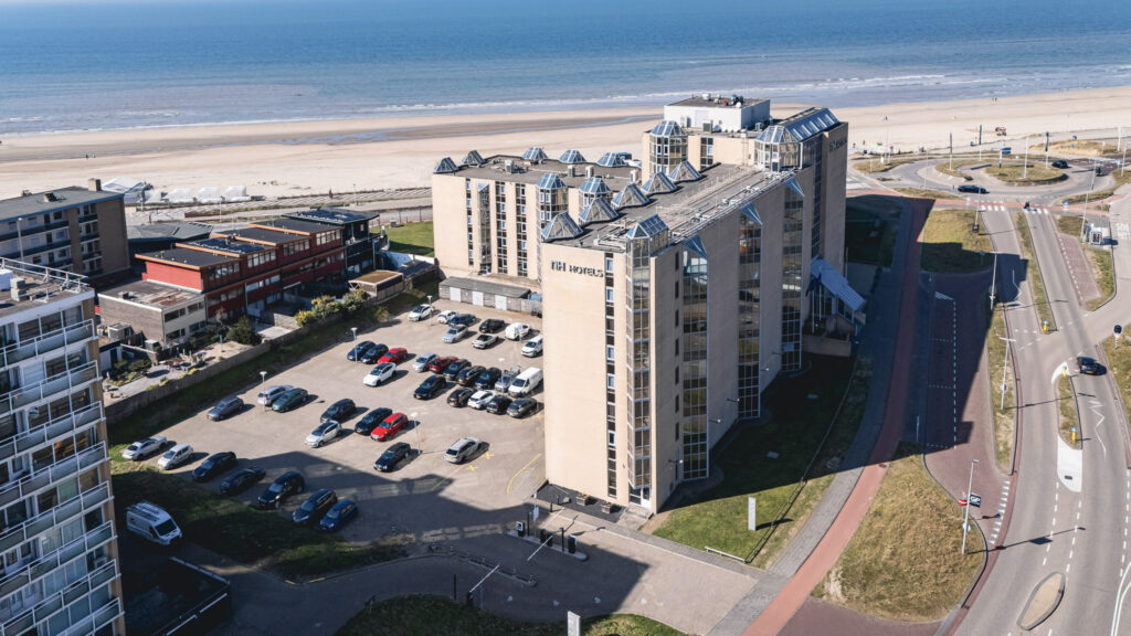 Aerial view of a seaside hotel with a parking lot full of cars, located near a sandy beach and ocean. Several buildings and roads surround the hotel, with clear skies and calm waves in the background.
