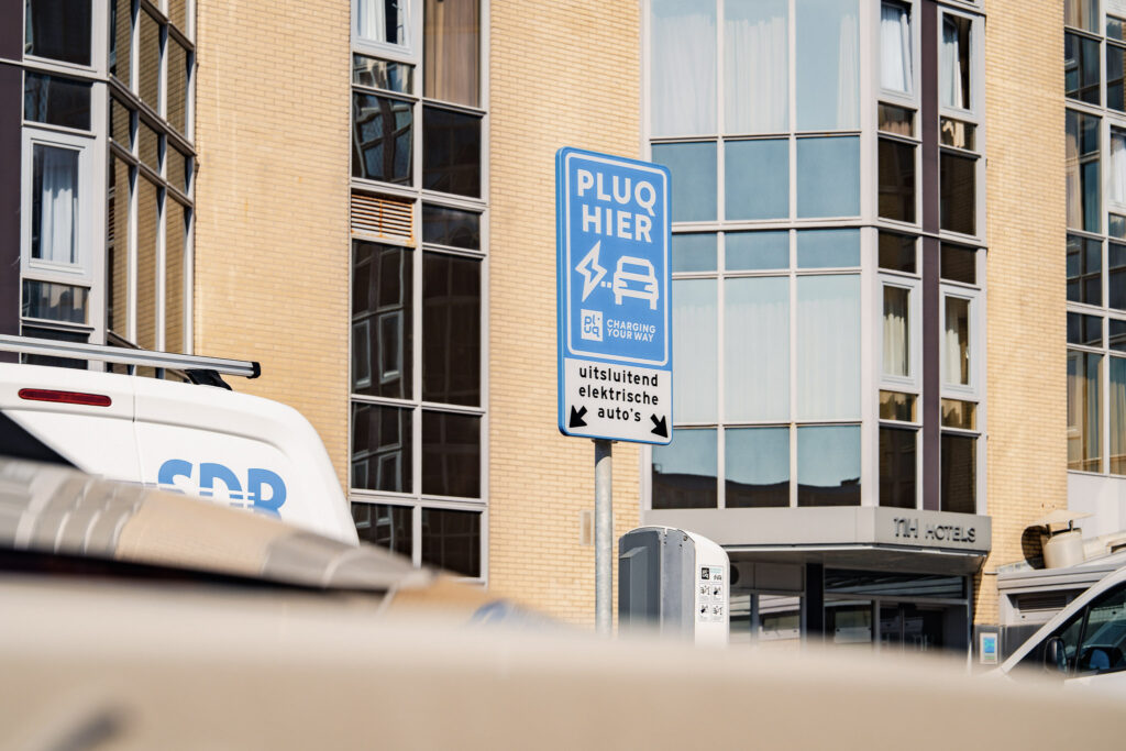 A blue sign in Dutch reads “PLUG HIER” and indicates a charging station for electric cars, set in front of a modern brick building with large windows. Several parked vehicles are visible nearby.
