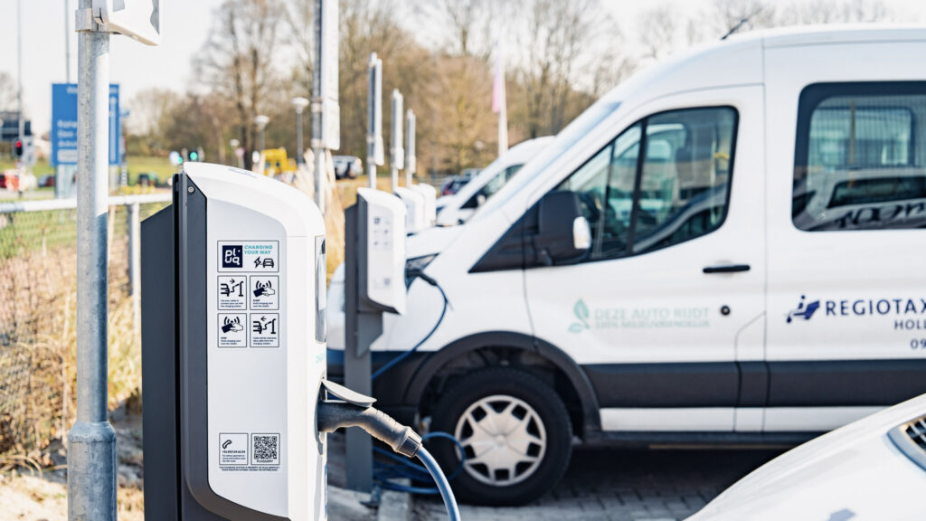An einem sonnigen Tag stehen Elektro-Lieferwagen auf einem Außenparkplatz in einer Reihe von Ladestationen und sind an diese angeschlossen; an der nächstgelegenen Ladestation sind Beschriftungen und Anweisungen zu sehen. Im Hintergrund sind Bäume und Schilder zu erkennen.