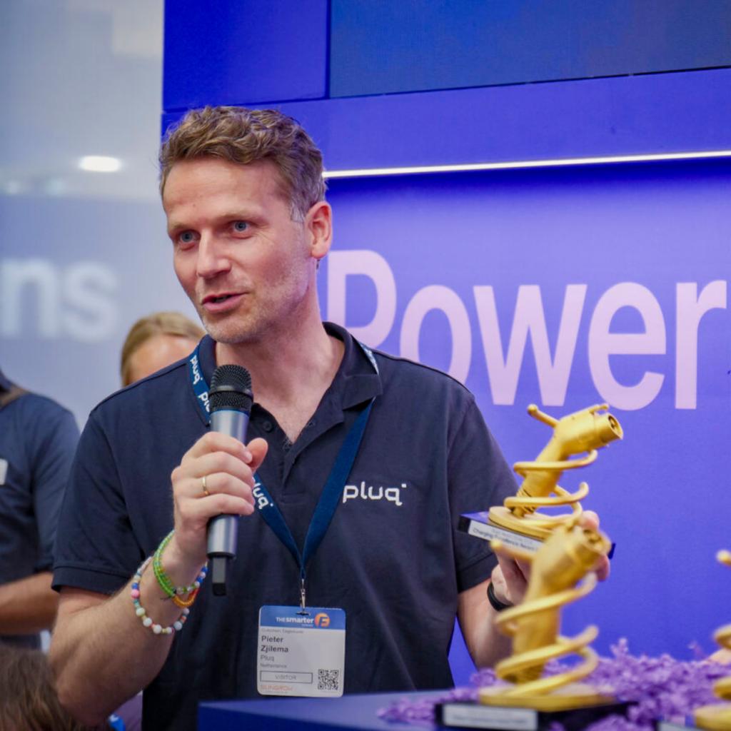 A man holding a microphone and a yellow 3D-printed award stands in front of a blue Power sign at an event, wearing a pluq shirt and name badge.