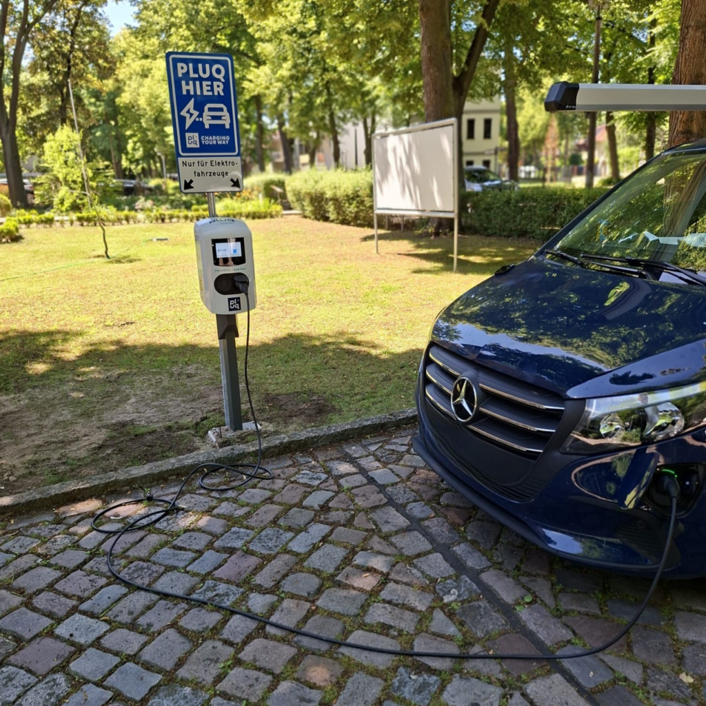 A blue Mercedes electric van is charging at a public charging station on a cobblestone street, with a PLUG HIER sign and trees in the background.